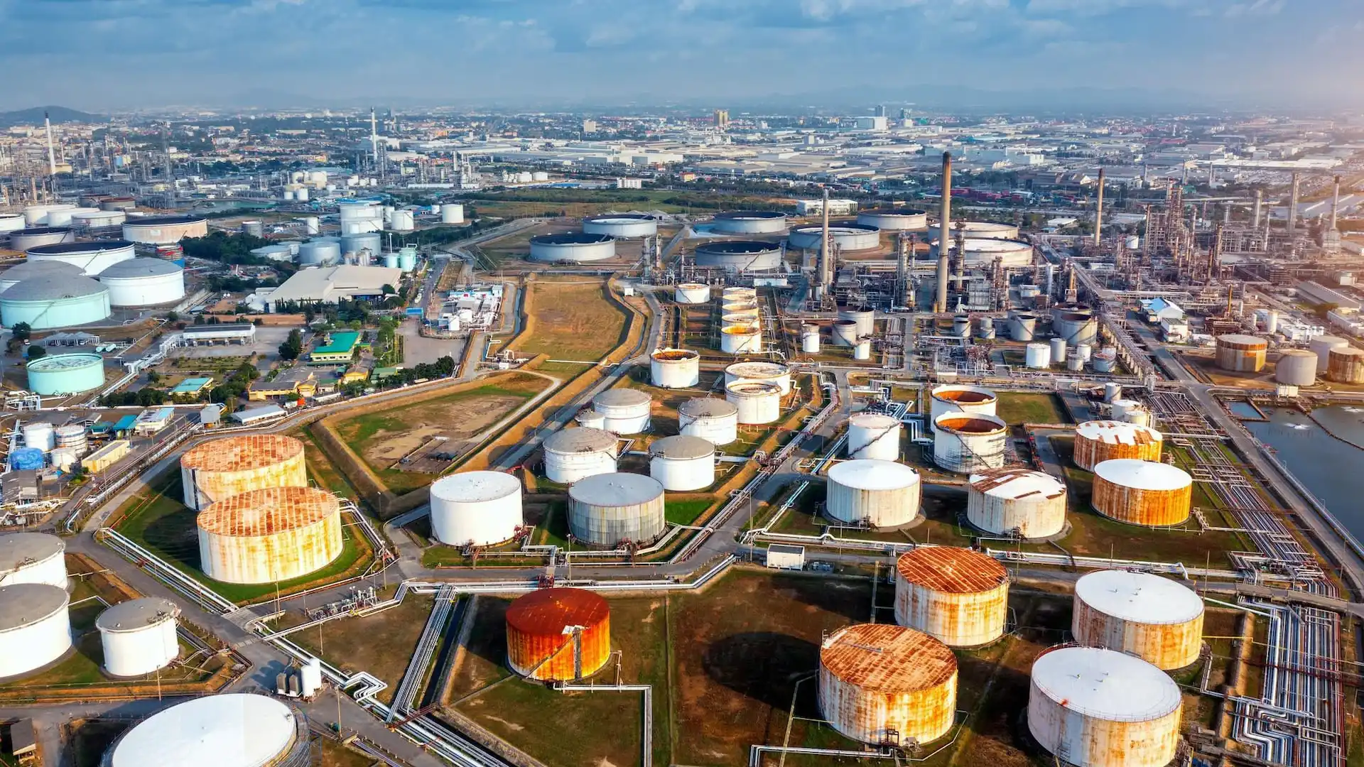 Aerial view of a large industrial oil and gas refinery and storage complex located near an urban area. Numerous cylindrical storage tanks, processing units, pipelines, and transport infrastructure are spread across the site in a dense network. Tanks of varying sizes are connected by roads and pipe systems, while additional industrial facilities and city buildings are visible in the background. The image illustrates large-scale energy production, storage, and distribution infrastructure.