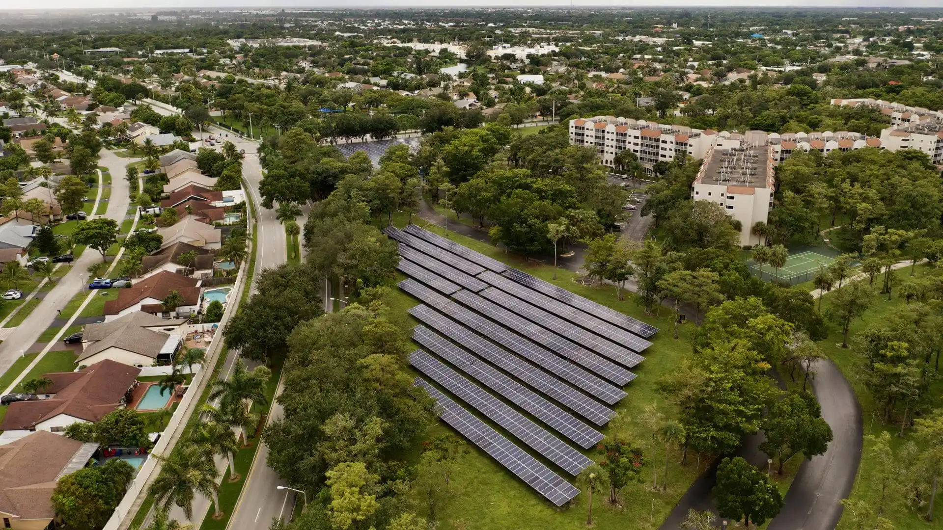 Aerial view of a ground-mounted solar farm installed on a grassy, tree-lined site within a suburban neighborhood. Multiple rows of solar panels are arranged in parallel between residential streets and apartment buildings. Dense greenery surrounds the installation, while flooded areas and water-covered land are visible in the distance, highlighting the contrast between renewable energy infrastructure and the surrounding urban landscape.