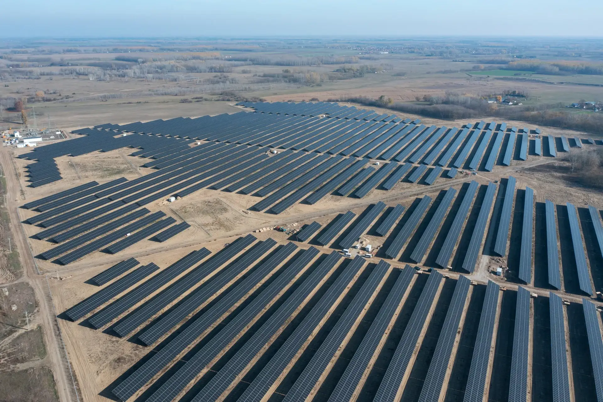 Aerial view of a large utility-scale solar power plant covering a vast open landscape. Hundreds of long, parallel rows of dark solar panels stretch across a cleared site, forming a geometric pattern visible from above. Access roads divide the installation into sections, with small service structures scattered throughout. Agricultural fields and flat rural terrain surround the solar farm, illustrating large-scale renewable energy generation infrastructure.