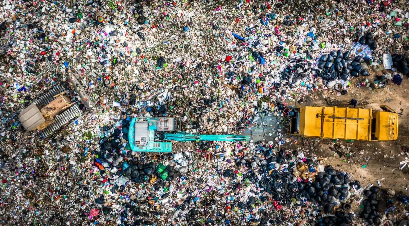 Aerial view of a large landfill site densely covered with mixed waste and discarded materials. A turquoise excavator operates in the center of the scene beside a yellow waste collection truck, surrounded by piles of plastic bags, packaging, and debris. The overhead perspective highlights the scale of waste accumulation and waste management operations, illustrating environmental challenges related to disposal, recycling, and resource recovery.